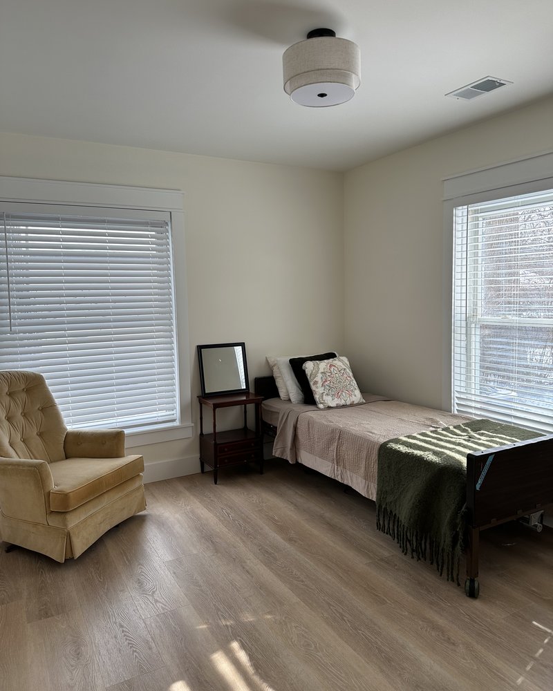 Resident bedroom with a tan tufted armchair, hospital bed, and sunny windows